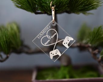 Op de afbeelding: Een transparante acryl sleutelhanger met het woord "GAMING" en twee videogame controllers die met een snoer verbonden zijn en een hartvorm vormen. De tekst "Name + Name" is in het hart gegraveerd.