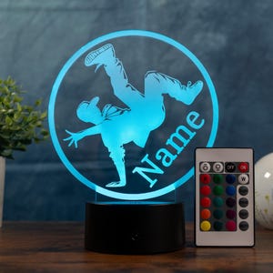 Könnte beinhalten: Eine blau beleuchtete Lampe zeigt eine Breakdance-Figur in einem kreisförmigen Rahmen mit dem Wort "Name". Die Lampe hat einen schwarzen Sockel, und eine Fernbedienung mit Farboptionen befindet sich in der Nähe. Die Lampe ist ein dekorativer Artikel.