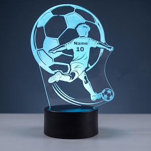 Op de afbeelding: Een lichtblauwe LED-lamp met een acryl uitgesneden figuur van een voetballer die tegen een bal schopt. De speler staat voor een grote voetbalafbeelding. De tekst "Name" en "10" staat op het shirt van de speler. De lamp heeft een zwarte voet.