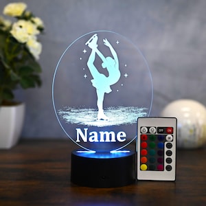 Könnte beinhalten: Eine weiße Silhouette einer Person, die Schlittschuh läuft, auf blauem Hintergrund mit Sternen. Der Hintergrund ist ein Kreis mit dem Wort "Name" darunter. Die Lampe steht auf einem schwarzen Sockel mit einer Fernbedienung rechts. Die Fernbedienung hat verschiedene Farboptionen.
