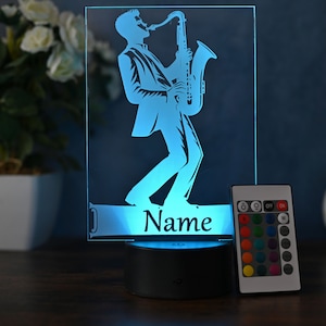 Op de afbeelding: Een blauw 3D-acryl nachtlampje met een silhouet van een persoon die saxofoon speelt. Het lampje heeft een afstandsbediening met meerdere kleur opties en een voet met het woord "Naam" erop.