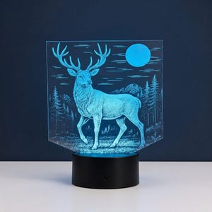 Könnte beinhalten: Eine flache, rechteckige Acryllampe mit einer geätzten Illustration eines Hirsches, der in einem Wald steht, mit einem Vollmond am Himmel. Das Design leuchtet in hellem Blau und ist auf einem runden, schwarzen Kunststoffsockel montiert.