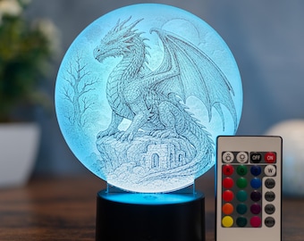 Op de afbeelding: Een blauw getinte drakenlamp met een gedetailleerd drakenontwerp geëtst in een cirkelvormig glaspaneel. De lamp staat op een zwarte voet en bevat een afstandsbediening met kleuropties. De draak zit op een rots met een kasteel op de achtergrond.