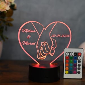 Peut inclure: Une veilleuse LED en forme de cœur avec deux mains qui se tiennent. La lumière est personnalisée avec les noms "Melina & Marcel" et la date "25.09.2024". La lumière est livrée avec une télécommande qui vous permet de changer la couleur de la lumière.