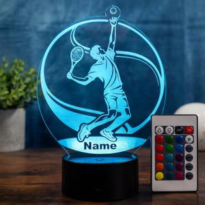Op de afbeelding: Een blauwe LED-lamp met een tennis thema, met een zwarte basis en een afstandsbediening. De lamp toont een silhouet van een tennisser in volle swing, met het woord "Name" aan de basis. De afstandsbediening heeft meerdere kleuropties.