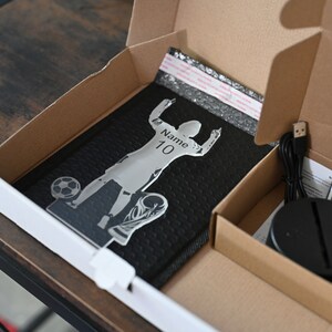 May include: A cardboard box with a black and silver 3D acrylic lamp inside. The lamp is a silhouette of a soccer player with the text "Name: 10" on the jersey. The box also contains a black base, a USB cable, and a remote control.