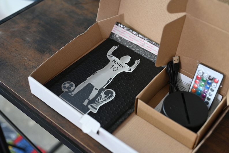 May include: A cardboard box with a black and silver 3D acrylic lamp inside. The lamp is a silhouette of a soccer player with the text "Name: 10" on the jersey. The box also contains a black base, a USB cable, and a remote control.
