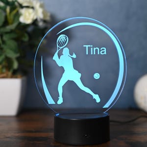 May include: A blue and white personalized LED night light with a silhouette of a tennis player holding a racket and a tennis ball. The name "Tina" is printed on the light.