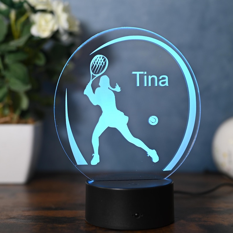 May include: A blue and white personalized LED night light with a silhouette of a tennis player holding a racket and a tennis ball. The name "Tina" is printed on the light.