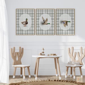 May include: Three framed duck prints with a gray plaid background and oval center. The ducks are depicted in various poses, with brown, green, and blue feathers. The prints are above a small white table and two wooden chairs.