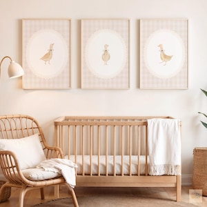 Op de afbeelding: Drie ingelijste prints met ganzenillustraties, elk tegen een beige en witte ruitjesachtergrond en een witte, op kant lijkende ovale rand. De prints hangen boven een houten ledikant in een kinderkamer.