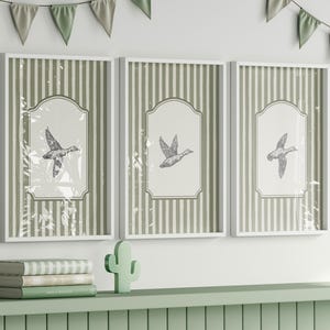 Peut inclure: Trois impressions encadr&eacute;es avec des cadres blancs, chacune avec une illustration d&eacute;taill&eacute;e d'un canard en vol. Le fond pr&eacute;sente des rayures verticales vertes et blanches. Une guirlande verte et blanche est suspendue au-dessus.