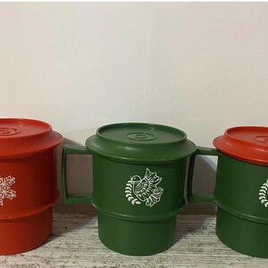 May include: Three vintage plastic mugs with lids. One is red with a white snowflake design, and two are green with a white bird and floral design. Each mug has a handle and a matching lid.