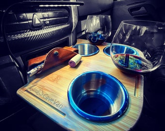 May include: A bamboo wine table with two stainless steel cup holders and two stemless wine glasses. A leather wine opener case and a cork are also on the table. The table is in a vehicle.