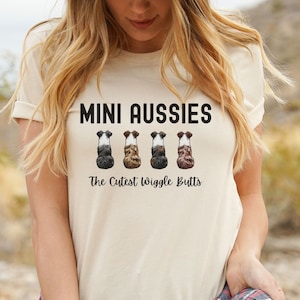 May include: A beige t-shirt with the words "MINI AUSSIES" in black, above an illustration of four dog rears. Below the illustration, the text reads "The Cutest Wiggle Butts". The shirt is worn by a person.
