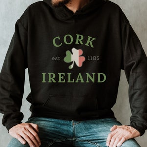 May include: Black hooded sweatshirt with the text "CORK IRELAND" in green, along with a shamrock design incorporating the Irish flag. The year "1185" is also displayed. The sweatshirt is worn by a person.