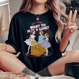K&ouml;nnte beinhalten: Schwarzes T-Shirt mit einem Motiv von drei M&auml;dchen, die ein Spiel mit einem Ouija-Brett spielen. Der Text auf dem Shirt lautet "Do you want to play a game?"
