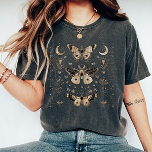 May include: A dark gray t-shirt with a gold and white moth design. The moth is surrounded by small flowers and stars. The shirt has a distressed look.