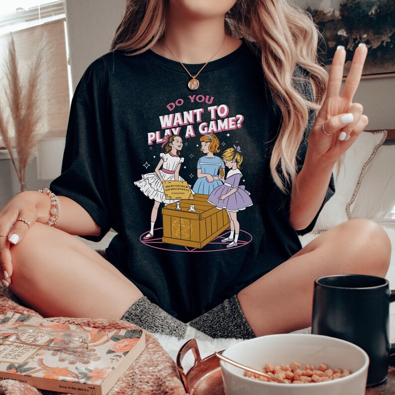 K&ouml;nnte beinhalten: Schwarzes T-Shirt mit einem Motiv von drei M&auml;dchen, die ein Spiel mit einem Ouija-Brett spielen. Der Text auf dem Shirt lautet "Do you want to play a game?"