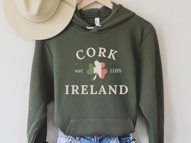 Op de afbeelding: Olijfgroene hoodie met de tekst "CORK IRELAND" en een klaverontwerp met de kleuren van de Ierse vlag. De hoodie heeft een capuchon met trekkoord en een voorzak. Een beige hoed hangt op de achtergrond aan een houten hanger.
