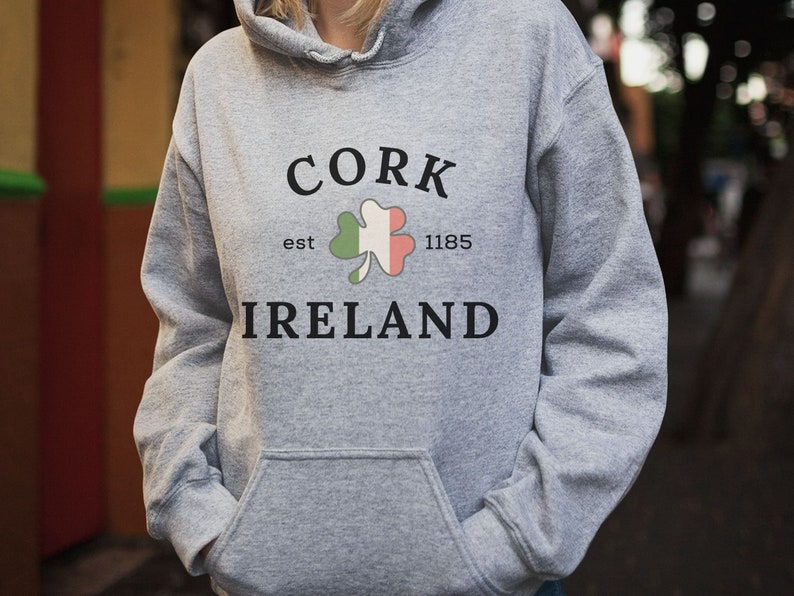 Op de afbeelding: Een grijze hoodie met een voorzak. De voorkant van de hoodie toont het woord "CORK" boven een klaverblad met de kleuren van de Ierse vlag en het woord "IRELAND" eronder. De tekst "est 1185" is ook aanwezig.