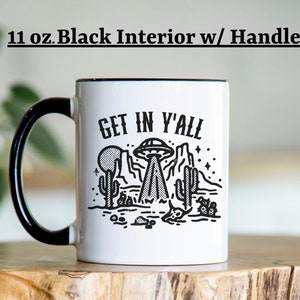 May include: White ceramic mug with a black handle and black rim. The mug has a black and white graphic of a UFO over a desert landscape with the text "Get in y'all".
