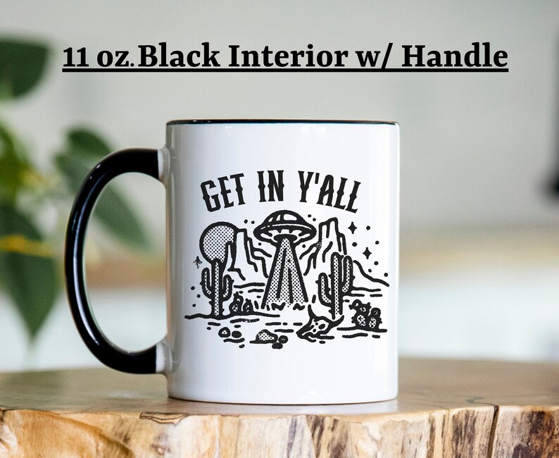 May include: White ceramic mug with a black handle and black rim. The mug has a black and white graphic of a UFO over a desert landscape with the text "Get in y'all".