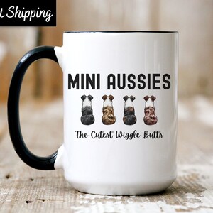 May include: White ceramic mug with a black handle and rim. The mug features the text "MINI AUSSIES" above four watercolor illustrations of dog rears. Below the text is "The Cutest Wiggle Butts".