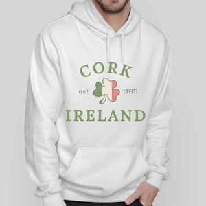 Op de afbeelding: Witte hoodie met een voorzak en een trekkoord. De hoodie heeft de tekst "CORK IRELAND" in groen, een klaverontwerp en het jaar "1185". Het ontwerp is gecentreerd.