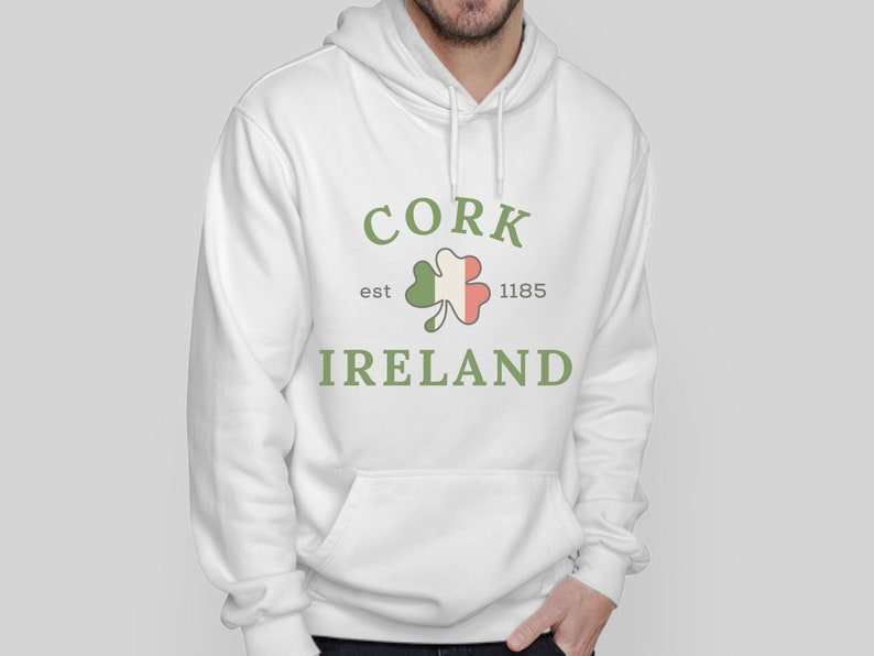 Op de afbeelding: Witte hoodie met een voorzak en een trekkoord. De hoodie heeft de tekst "CORK IRELAND" in groen, een klaverontwerp en het jaar "1185". Het ontwerp is gecentreerd.