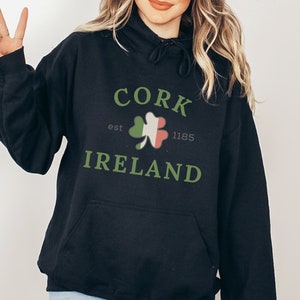 Op de afbeelding: Zwarte hoodie met de tekst "CORK IRELAND" in groen, met een klaverontwerp. De hoodie heeft ook de tekst "est 1185".