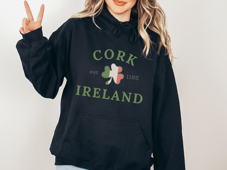 Op de afbeelding: Zwarte hoodie met de tekst "CORK IRELAND" in groen, met een klaverontwerp. De hoodie heeft ook de tekst "est 1185".