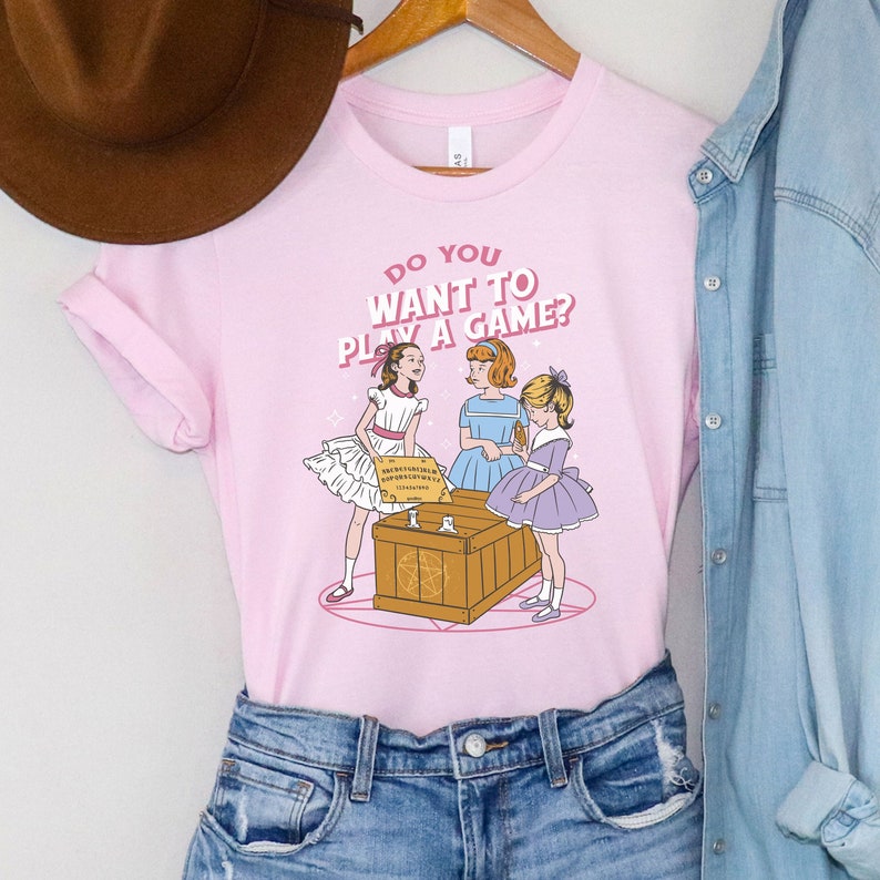 K&ouml;nnte beinhalten: Rosa T-Shirt mit einem Motiv von drei M&auml;dchen in Kleidern, die um eine Holzkiste mit einem Ouija-Brett darauf stehen. Der Text auf dem T-Shirt lautet "Do you want to play a game?"