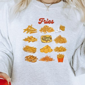 Pu&ograve; includere: Una felpa grigia con la parola "Fries" in lettere rosse e una variet&agrave; di illustrazioni di patatine fritte. Le illustrazioni includono patatine fritte ricce, patatine fritte a waffle e patatine fritte francesi.