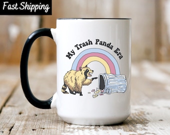 May include: White ceramic mug with a black handle and rim. The mug features a cartoon raccoon and a tipped-over trash can with the text "My Trash Panda Era" above a rainbow design. The mug has a capacity of approximately 15 ounces.