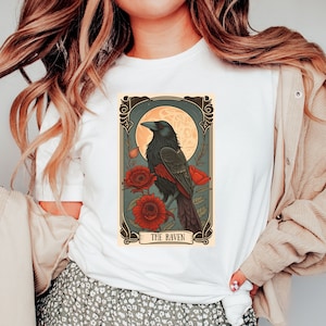 May include: A white t-shirt with a vintage-style illustration of a raven perched on a branch with red flowers. The illustration is framed with a decorative border and the text "THE RAVEN" is printed below the image.