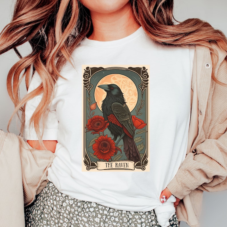May include: A white t-shirt with a vintage-style illustration of a raven perched on a branch with red flowers. The illustration is framed with a decorative border and the text "THE RAVEN" is printed below the image.