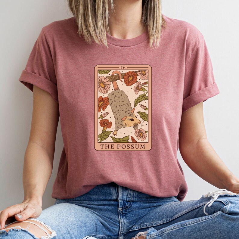 May include: A pink t-shirt featuring a tarot card graphic of a possum hanging from a branch with flowers. The card reads "The Possum" and "IV".