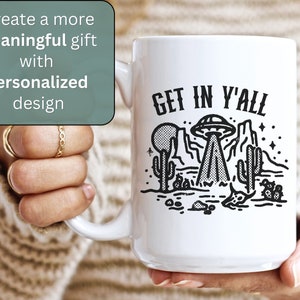 May include: White ceramic mug with a black and white illustration of a UFO over a desert landscape. The text "GET IN Y'ALL" is printed on the mug.