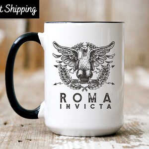 May include: White ceramic mug with a black handle and rim. The mug features a detailed black and white illustration of an eagle with outstretched wings, a laurel wreath, and the words "ROMA INVICTA". The text "Fast Shipping" is in a black box.