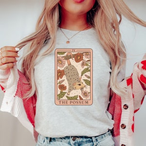 May include: A light grey t-shirt with a graphic of a tarot card featuring a possum hanging from a branch with flowers. The card is labelled "The Possum" and "IV".