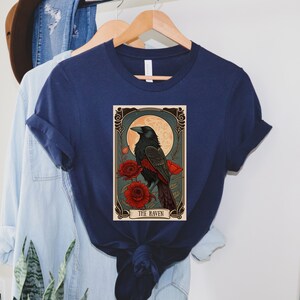 May include: A navy blue t-shirt with a vintage-style illustration of a raven perched on a branch with red flowers. The illustration is framed with a decorative border and includes the text "THE RAVEN".