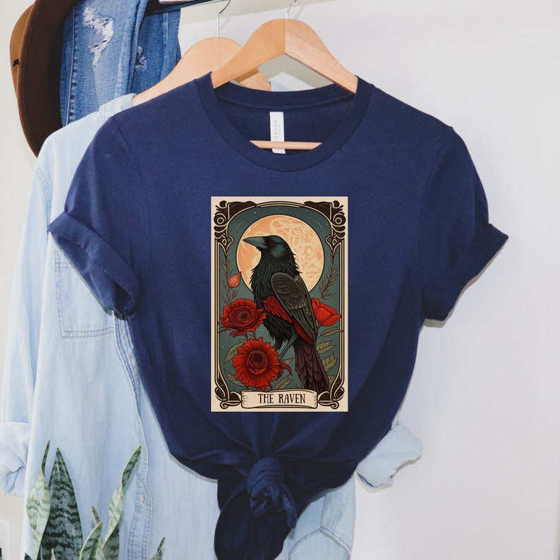 May include: A navy blue t-shirt with a vintage-style illustration of a raven perched on a branch with red flowers. The illustration is framed with a decorative border and includes the text "THE RAVEN".