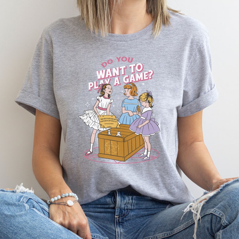 K&ouml;nnte beinhalten: Ein graues T-Shirt mit dem Text "Do you want to play a game?" und einer Illustration von drei M&auml;dchen in Kleidern, die um eine Holzkiste mit einem Pentagramm stehen.