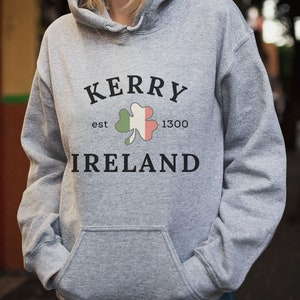 Op de afbeelding: Grijs hoodie met de tekst "Kerry est 1300 Ierland" en een groene klaver met oranje en witte accenten.