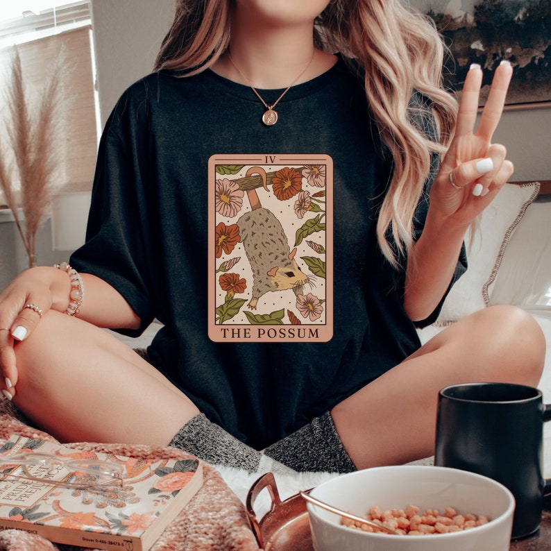 May include: A black t-shirt featuring a tarot card graphic with a possum hanging upside down. The card reads "The Possum" and "IV".