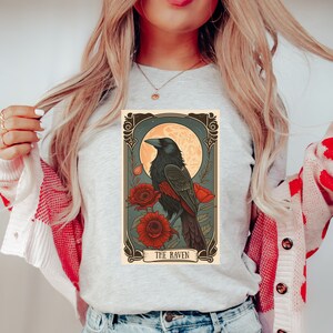 May include: A gray t-shirt with a vintage-style illustration of a raven perched on a branch with red flowers. The illustration is framed with a decorative border and the text "THE RAVEN" is at the bottom.