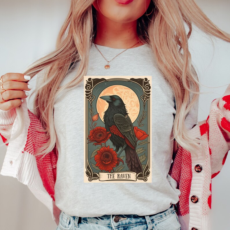May include: A gray t-shirt with a vintage-style illustration of a raven perched on a branch with red flowers. The illustration is framed with a decorative border and the text "THE RAVEN" is at the bottom.