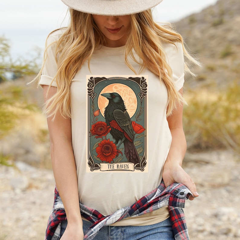 May include: A white t-shirt with a vintage-style illustration of a raven perched on a branch with red flowers. The illustration is framed with a gold border and the text "THE RAVEN" is printed below the image.