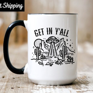 May include: White ceramic mug with a black rim and handle. The mug features a black and white illustration of a UFO over a desert landscape with the text "Get in Y'all".